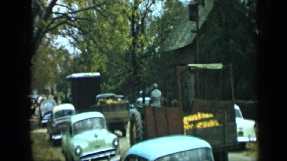 1959 Aledo, Texas home-movie frame showing vintage cars along a shaded street beside a loaded farm trailer.