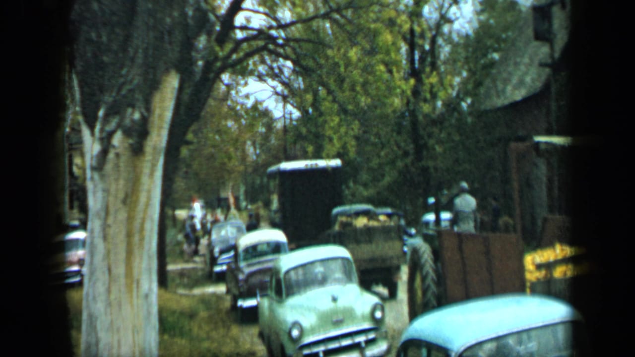 Vintage cars and people on a shaded road beside a trailer and trees.