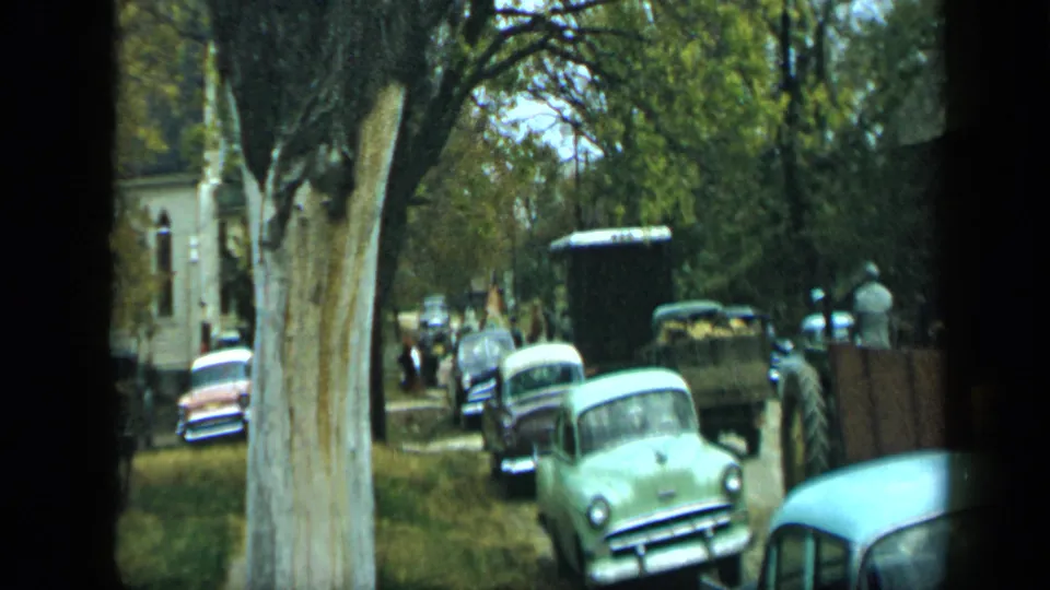 Aledo, Texas street scene from 1959 with a large roadside tree, classic cars, pedestrians, and a loaded trailer.