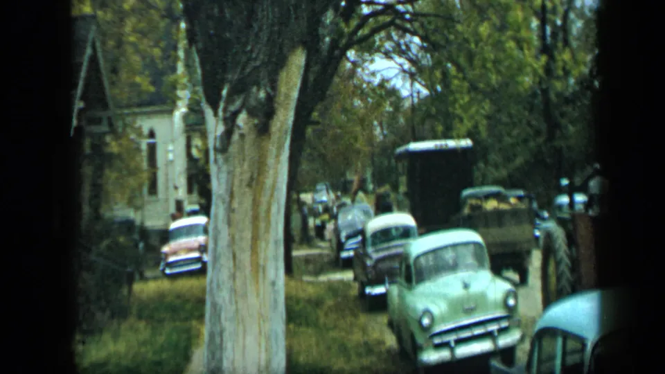 Final frame of the 1959 Aledo street view, with vintage automobiles, a tree trunk, pedestrians, and a farm trailer.