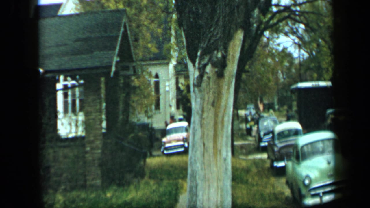Parked vintage cars stand beside trees and a brick house along a roadside.