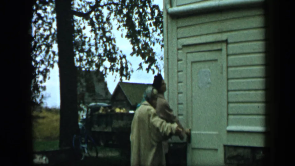 Aledo, Texas 1959 frame with an adult near a clapboard doorway, trees, parked vehicles, and a small church-like building in the background.