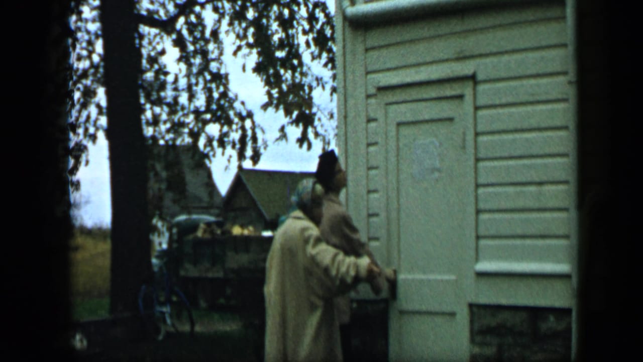 Two adults stand near a white house door under tree branches, with a bicycle and small shed in the background.