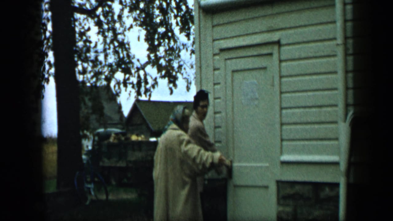 A woman in a light coat and headscarf and a man stand at the doorway, both turned partly back.