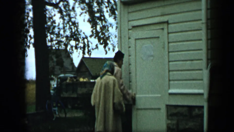 1959 Aledo, Texas 8mm frame with two adults nearing a white house door as the camera looks past dark foreground film edges.