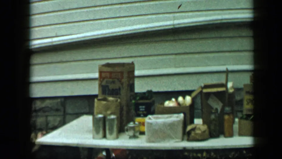 Aledo, Texas 1959 frame with boxed food, cans, eggs, bottles, and sacks on an outdoor table beside a house exterior.