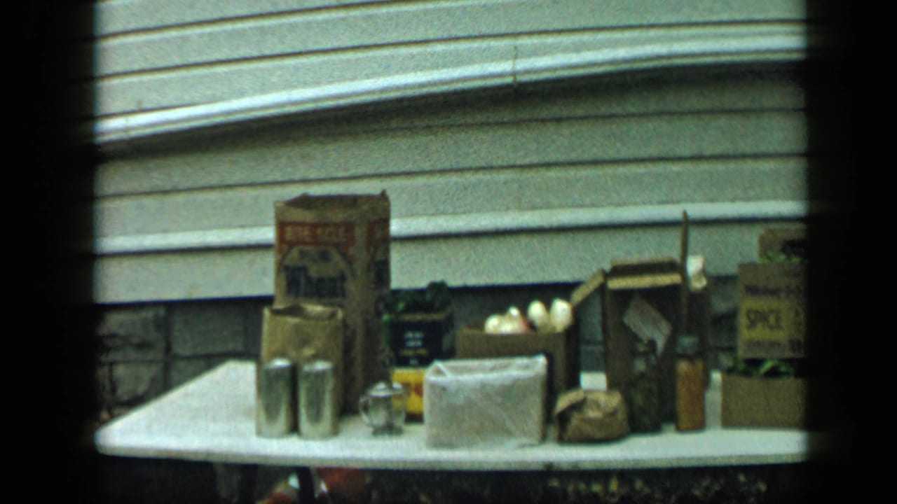 Outdoor table with flour box, metal canisters, paper bags, onions, and household goods against house siding.