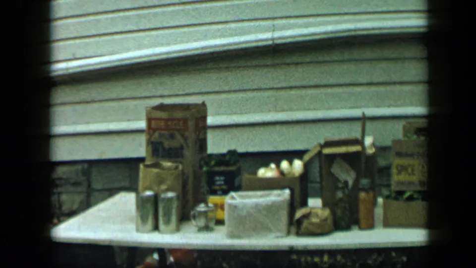 1959 Aledo, Texas home-movie frame showing a white outdoor table set with flour packaging, cans, bags, onions, and small household containers.