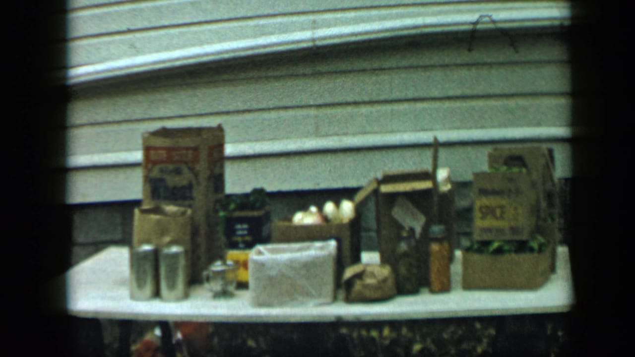 1959 Aledo, Texas home-movie frame of an outdoor table holding pantry goods, produce, metal canisters, and labeled packaging.