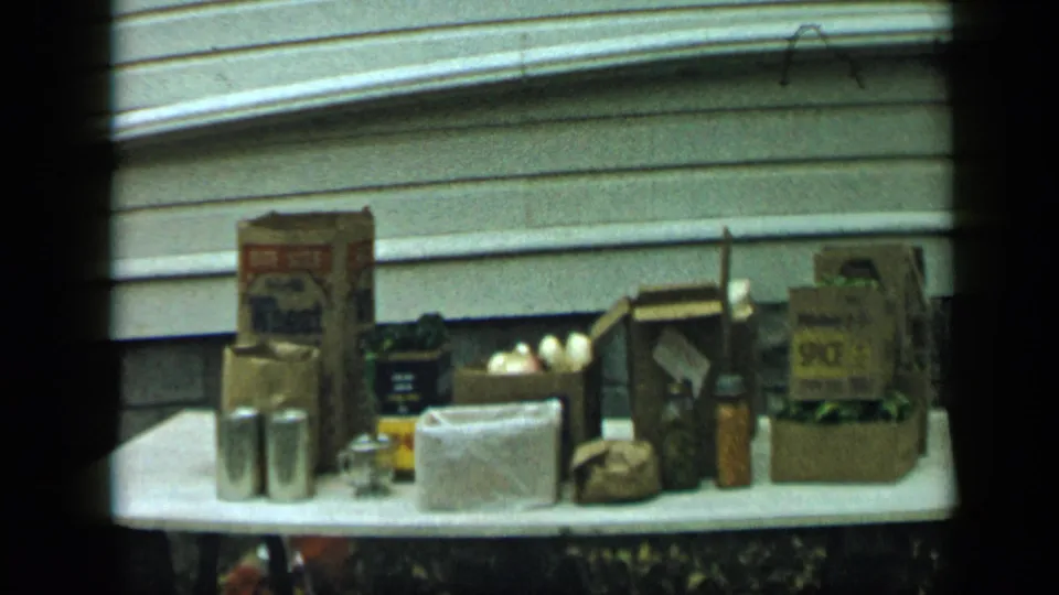 1959 Aledo, Texas home-movie frame of an outdoor table holding pantry goods, produce, metal canisters, and labeled packaging.