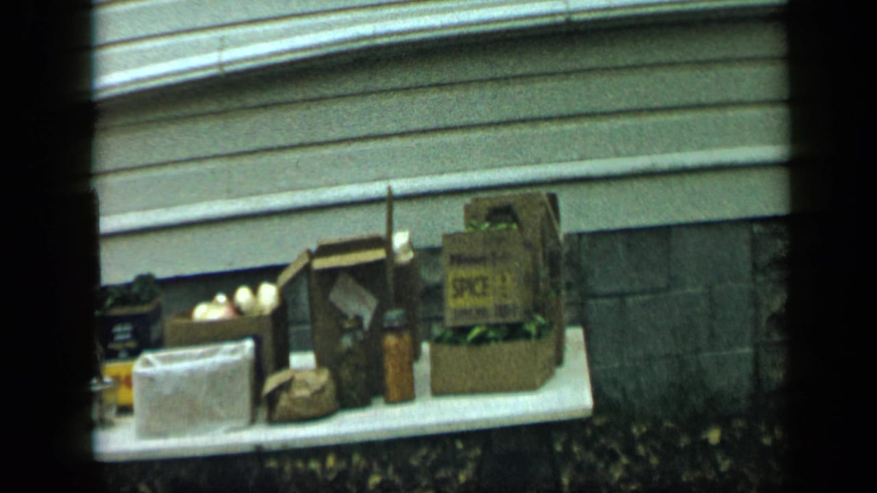 White outdoor table holding cardboard boxes and jars beside house siding and stone foundation.
