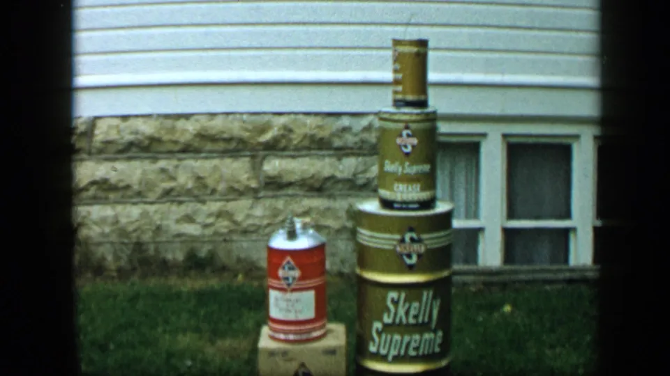 1959 Aledo, Texas frame showing stacked Skelly Supreme and Daily Supreme cans on a box in front of house siding and a stone foundation.