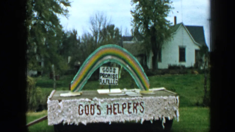 1959 Aledo, Texas frame of a homemade rainbow religious display on a lawn near houses and trees.