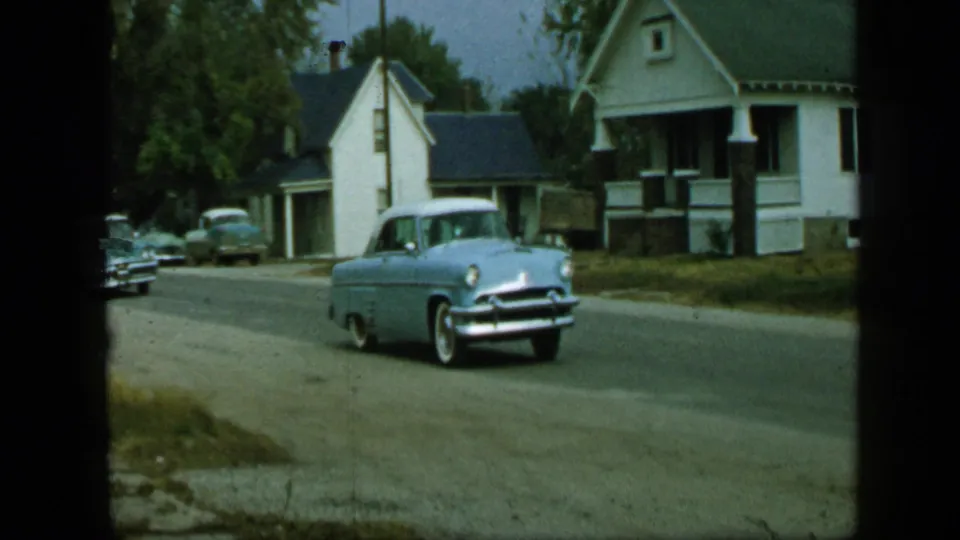 1959 Aledo, Texas frame showing a light blue mid-century car moving down a quiet residential street.