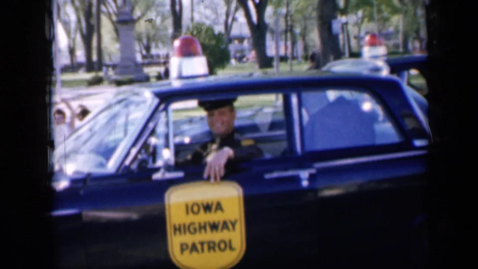 1959 Iowa 8mm frame showing an Iowa Highway Patrol officer in a dark patrol car, with a red roof beacon and yellow door sign visible.