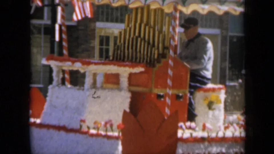 A 1959 Iowa parade float fills the frame, with an adult calliope player partly visible behind floral trim and American flags.