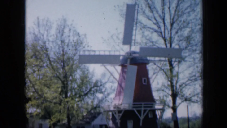 A Dutch-style red-and-white windmill stands among spring trees in Iowa, filmed in soft 8mm color.