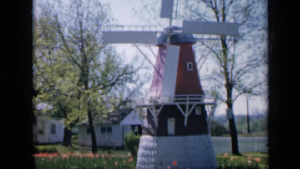 A red-and-white windmill stands in a landscaped Iowa setting with spring trees and tulips visible near the base.