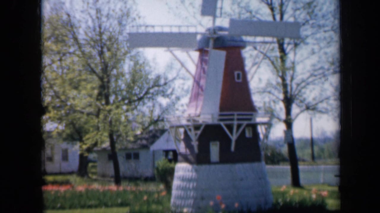 Small red-and-white windmill on a lawn with flowers, trees, and pale buildings behind it.