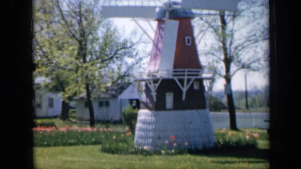 1959 Iowa home-movie frame of a small windmill with white trim, set among grass, flower beds, trees, and background buildings.