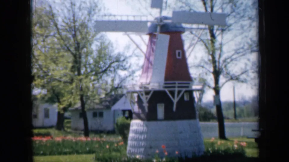 1959 Iowa 8mm view of the garden windmill, with its white base, red tower, trimmed lawn, flowers, and trees in soft film color.