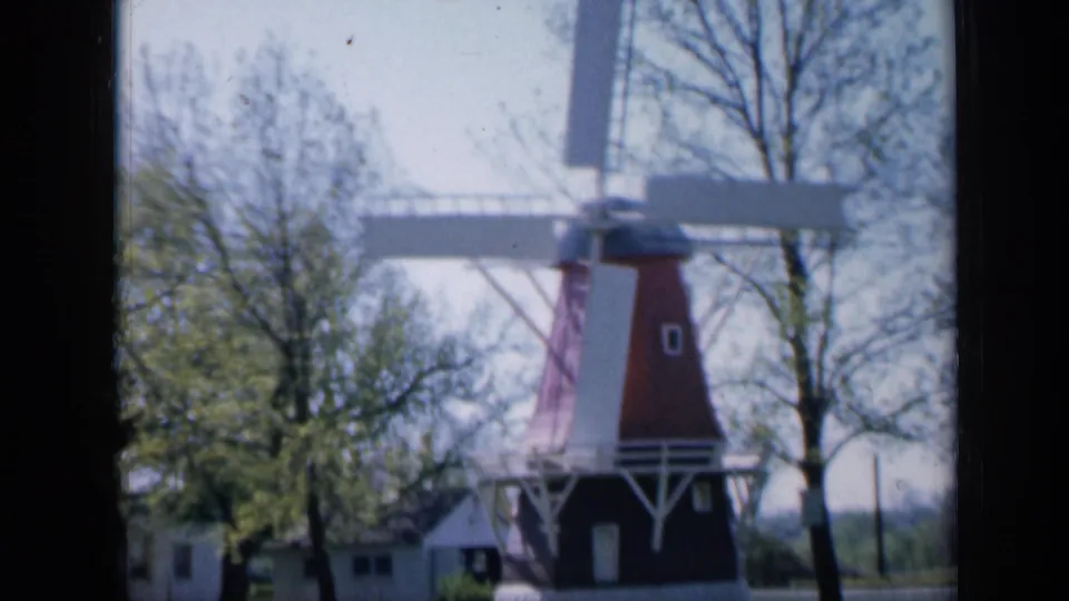 Soft 1959 Iowa 8mm frame showing the same small garden windmill in a landscaped outdoor setting with trees and flowers.