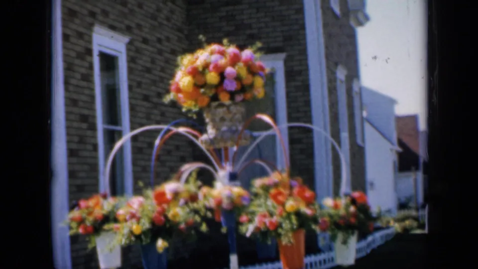 1959 Iowa 8mm frame showing orange, yellow, and pink flowers arranged before a brick house exterior.