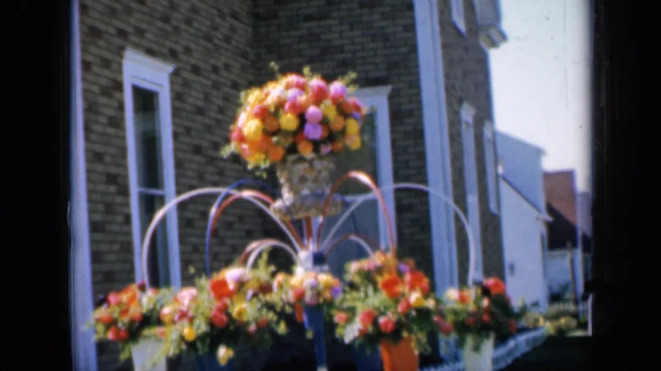 1959 Iowa 8mm frame showing hanging pots and a central floral urn outside a brick home.