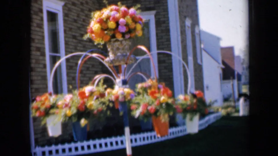 1959 Iowa 8mm frame showing a colorful floral display, white fence, and brick house exterior in daylight.