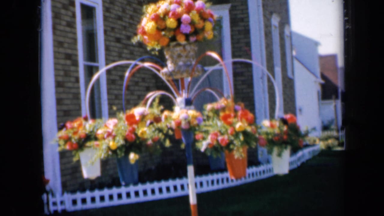 Front yard floral display with urn, hanging baskets, lawn, brick house, and white picket fence.