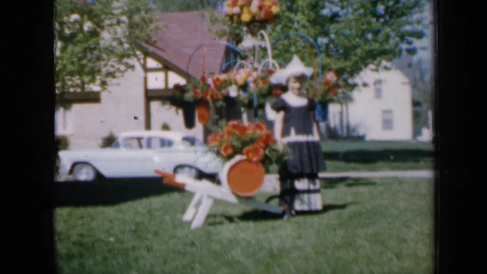 1959 Iowa home-movie frame showing a young female subject in a dark dress and white hat posing by a floral display on a suburban lawn.