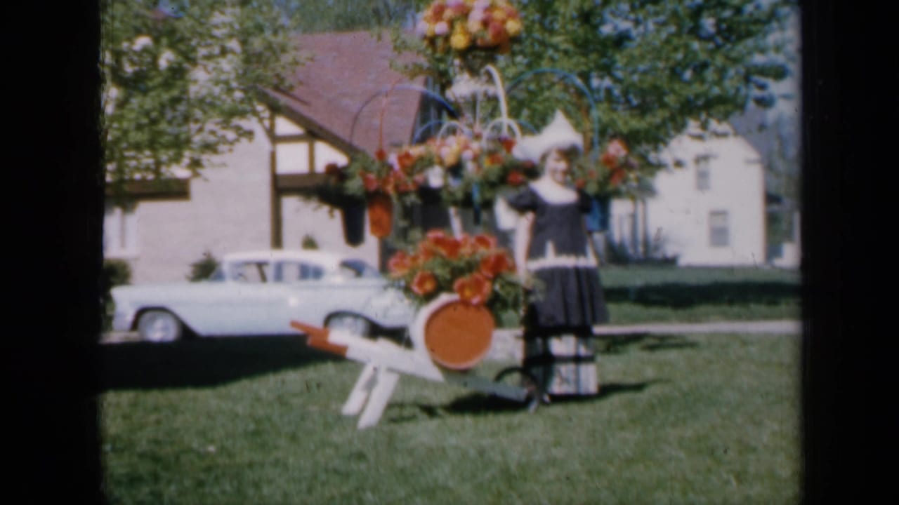 Girl in traditional dress posing beside a flower-covered cart on a suburban lawn.