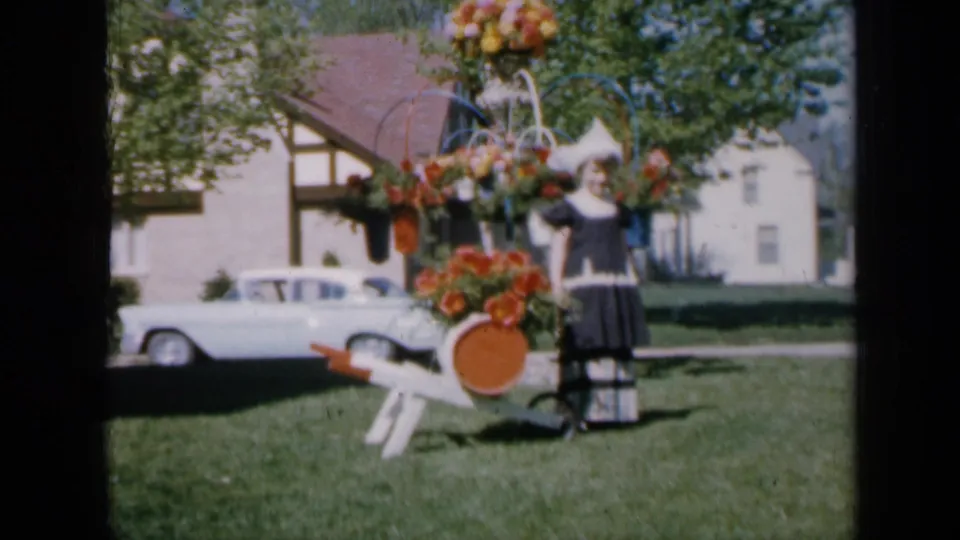 In 1959 Iowa home-movie footage, a girl in Dutch-style dress stands beside a decorated flower cart with a white sedan and house behind her.