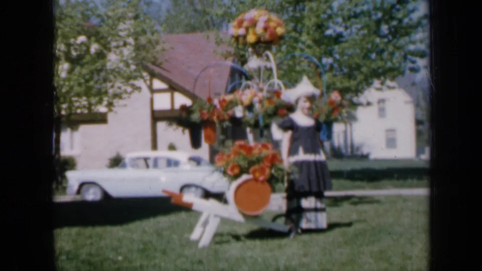 The girl remains posed next to the floral cart in a sunny residential yard, with a mid-century white sedan parked near the house.