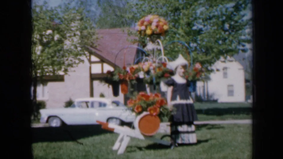A home-movie frame shows the girl beside the decorated cart, with flowers, lawn, a white sedan, and neighboring house details visible.