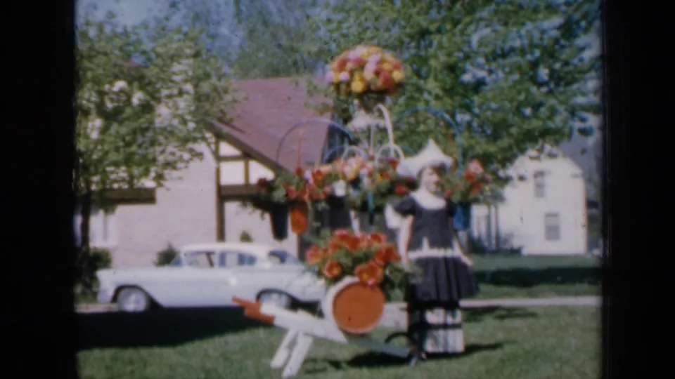The final frame holds on the girl and flower-decorated cart in an Iowa residential setting, with trees, house, and white sedan in view.