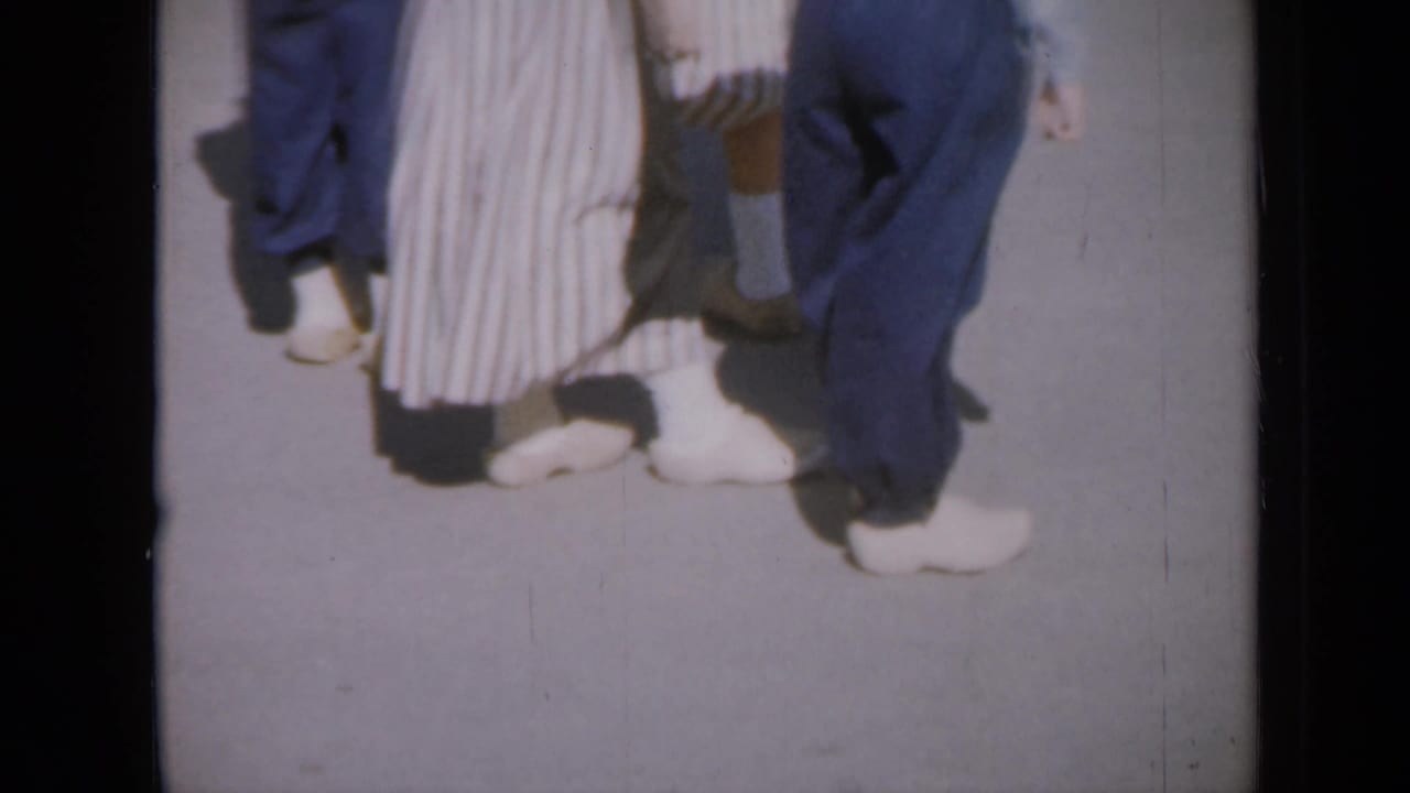 Lower legs of dancers in white shoes on a paved surface.