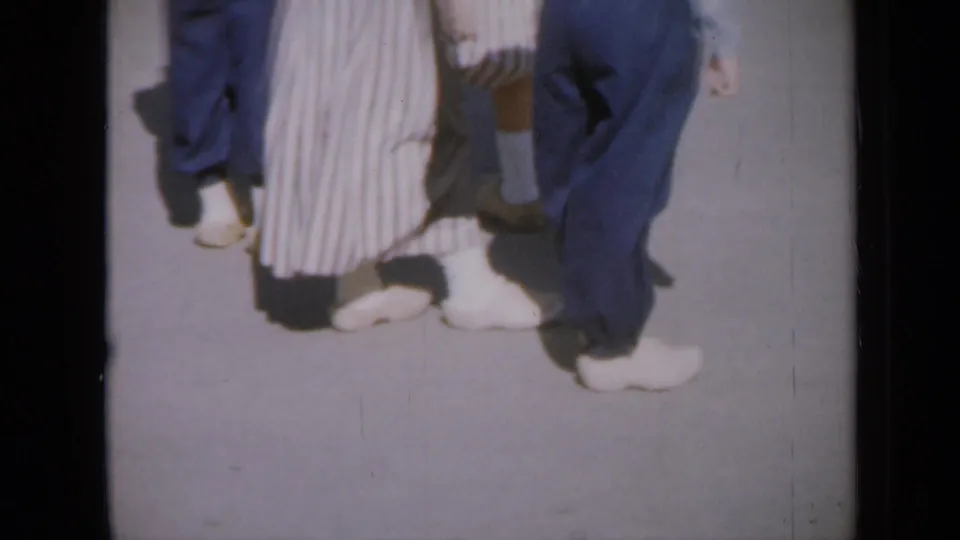 1959 Iowa home-movie frame showing dancers' lower legs, white shoes, blue pants, and a striped skirt on pavement.