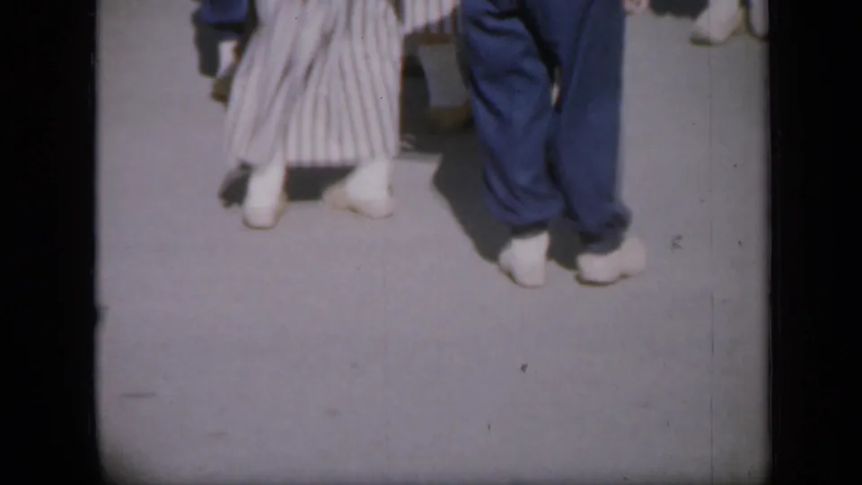 Low-angle 1959 Iowa frame with white dance shoes, blue trousers, and a striped skirt gathered on a paved area.