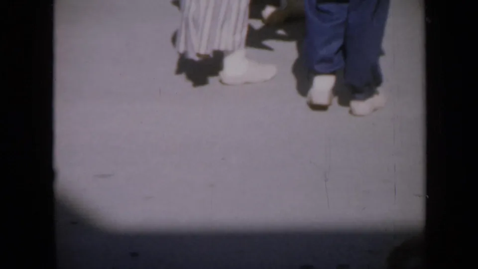 Home-movie view of dancers' feet in white shoes, with blue pants and a striped dress partly visible in bright sun.