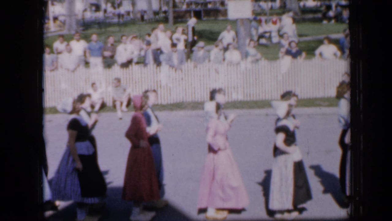 Costumed dancers walk past spectators behind a white picket fence on a sunny street.