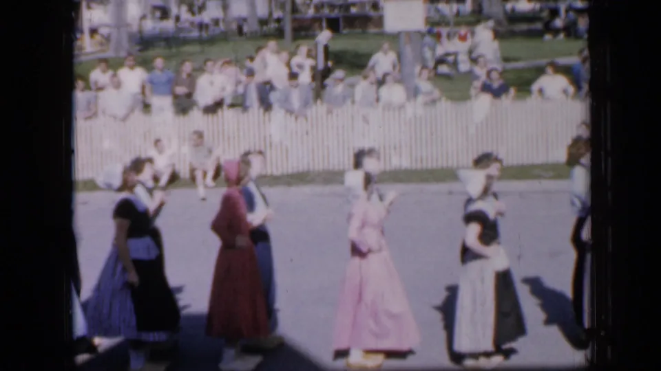 1959 Iowa home-movie frame of costumed dancers in long dresses moving past a crowd behind a white picket fence.