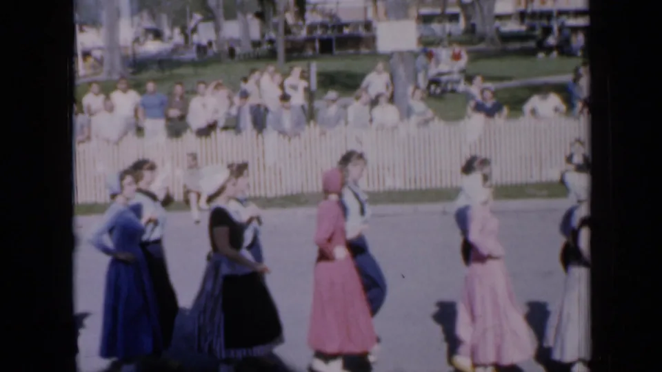 1959 Iowa frame showing folk dancers in dresses and shirts moving along a street before seated and standing spectators.