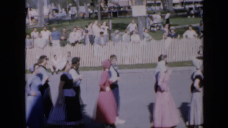 1959 Iowa home-movie view of dancers crossing the frame, with a white fence, spectators, grass, and cars in the background.