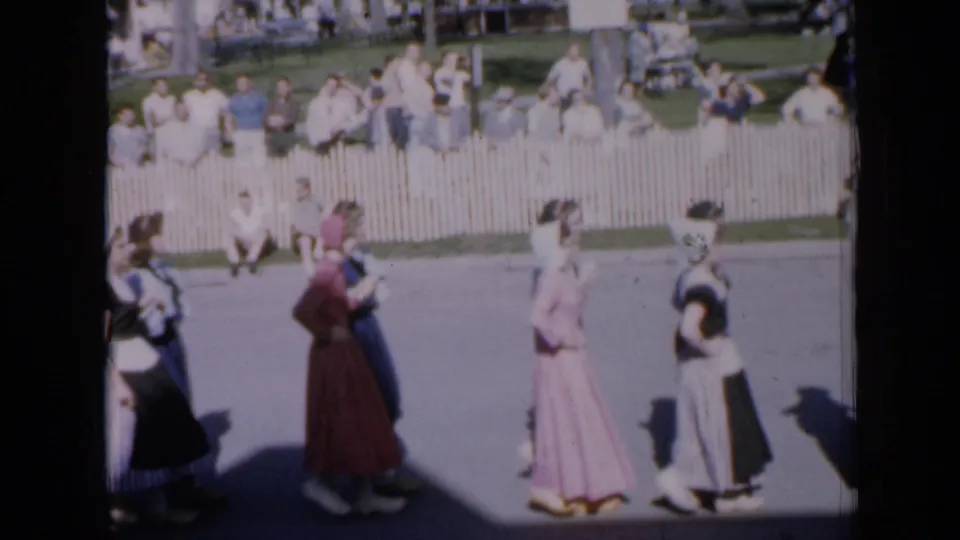 1959 Iowa frame of a costumed folk dance formation on a sunny street, seen from a soft 8mm home-movie scan.