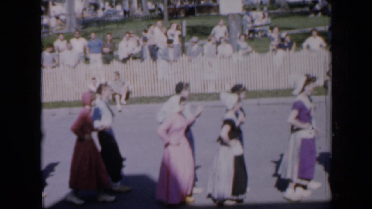 Dancers in long dresses and dark trousers perform near a white fence as spectators watch.