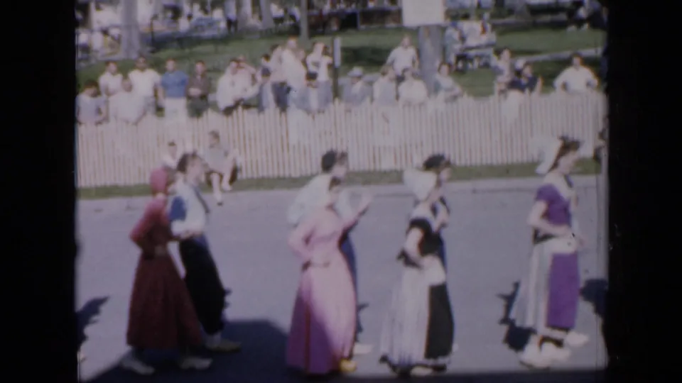 In Iowa 1959 home-movie footage, costumed dancers perform in a sunny park before spectators behind a white picket fence.