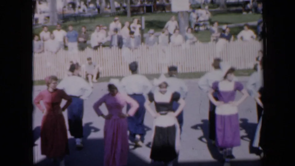 A folk-dance group in 1959 Iowa appears mid-routine, with women in long dresses and men in white shirts facing spectators.