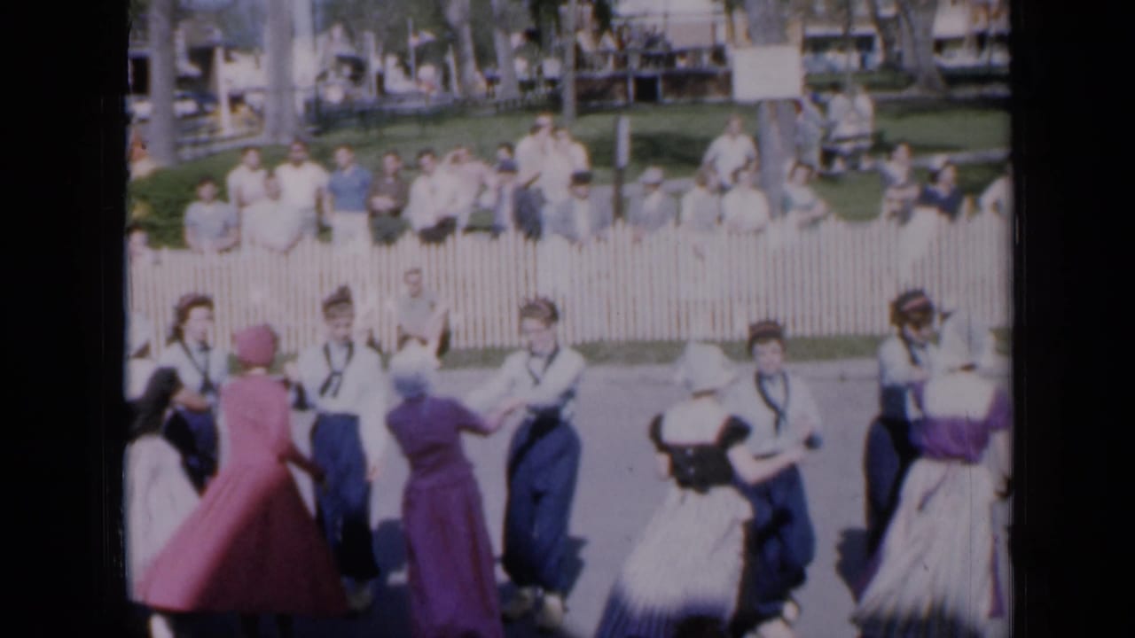 Costumed dancers perform in front of spectators behind a white picket fence.