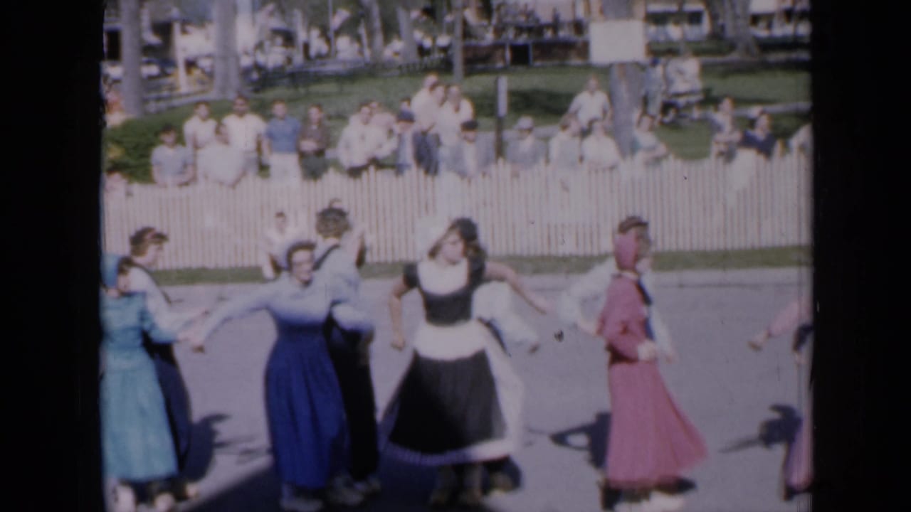 1959 Iowa 8mm frame of folk dancers in skirts, bonnets, shirts, and trousers performing before a seated park audience.